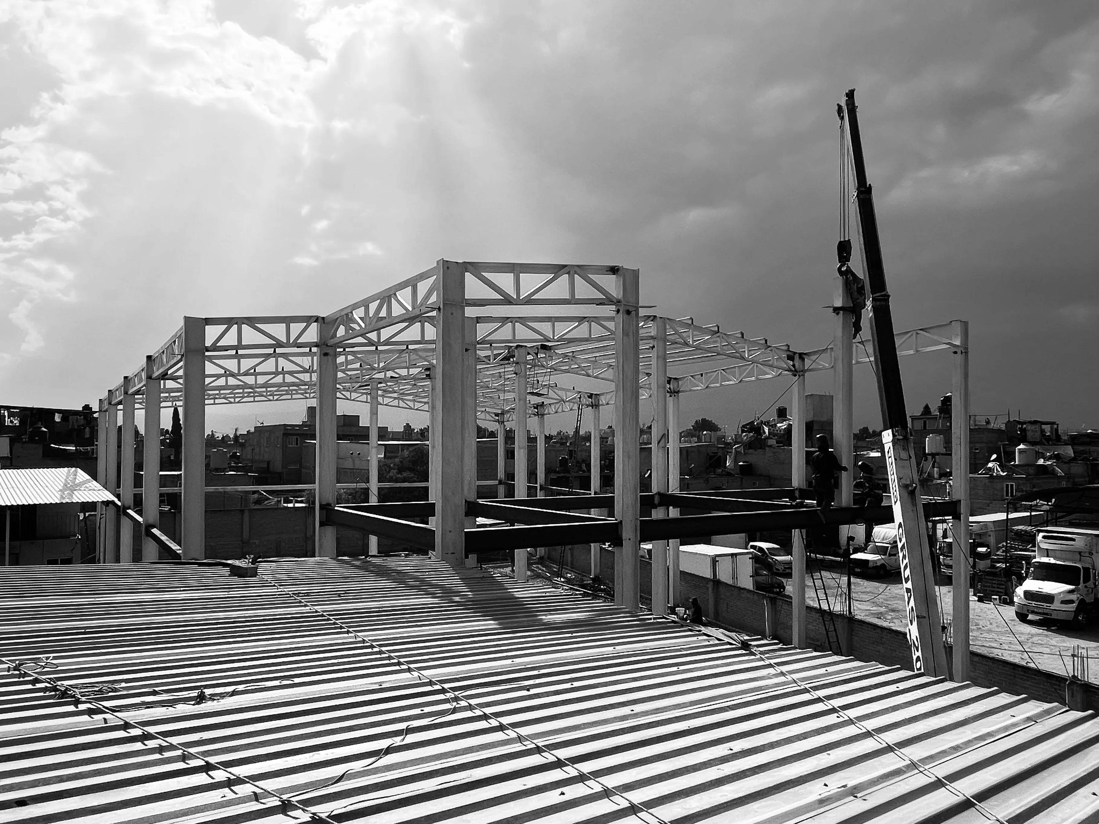 Estructura de nave industrial — B&W con rayos de sol entre nubes, vista desde la techumbre.