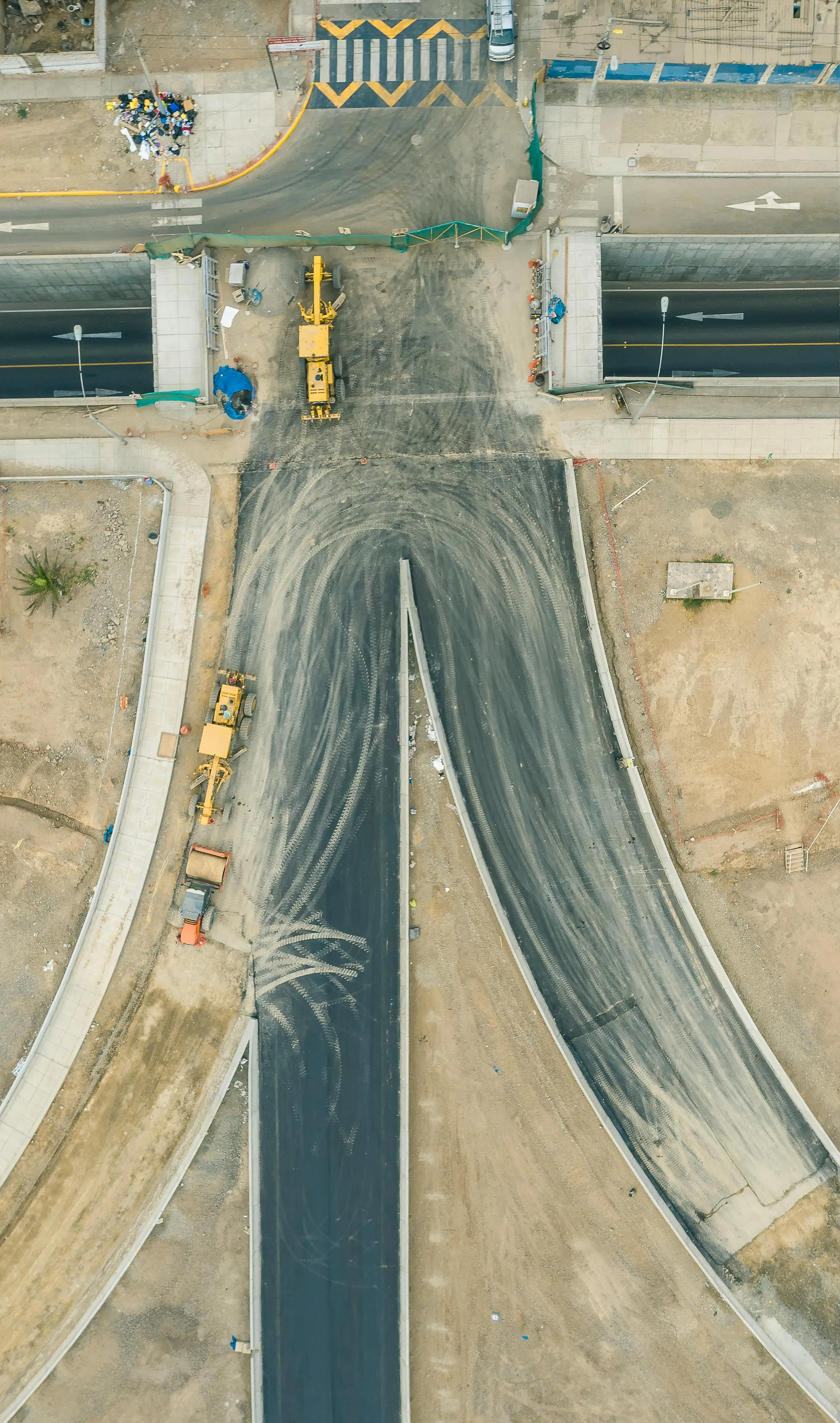 Vista cenital de obra vial: trazado geométrico de pavimento con maquinaria y cruce de calles.
